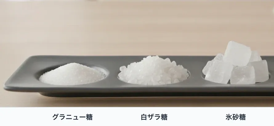 濃い灰色の仕切りのある陶板に、精製糖3種を盛って結晶の大きさを比べる。左からグラニュー糖(細かくきらめく粒)、白ザラ糖(粒のはっきり見える透明な結晶)、氷砂糖(角ばった透明な塊)。上白糖はグラニュー糖と、粉砂糖は写真の粒の細かさでグラニュー糖と見分けにくいため外した